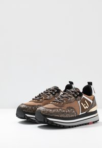 Zapatillas deportivas de malla marrón con detalles en negro, diseño con patrón, herrajes dorados y una suela gruesa negra con detalles en blanco y rojo.