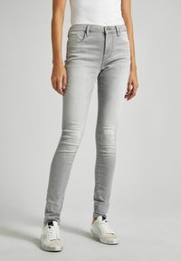 Jeans skinny desgastados en gris con diseño de cinco bolsillos, ligero desgaste y cierre de botón en la parte delantera, combinados con zapatillas blancas.