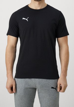 Mężczyzna w czarnej koszulce Puma i szarych spodniach dresowych Puma, stoi z rękami swobodnie opuszczonymi wzdłuż ciała.