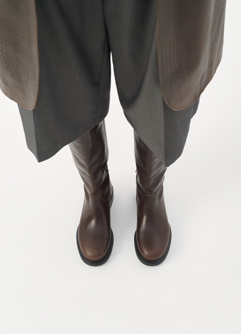 Bottes en cuir marron hauteur genou avec une texture lisse, bout arrondi et un léger talon, associées à un pantalon en tissu gris.