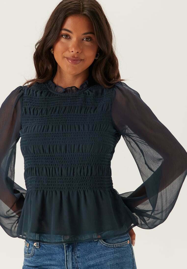 Tmavě modrá blůza s nabíranou texturou, průhlednými nadýchanými rukávy a volánkovým výstřihem. Peplum spodní část přidává objem. Nosit s džínovými šortkami.