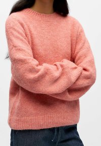 Šviesiai rožinis megztinis su apvalia iškirpte, nuleistais pečiais ir ribbingu apačioje. Medžiaga atrodo minkšta ir šiek tiek pūkuota.