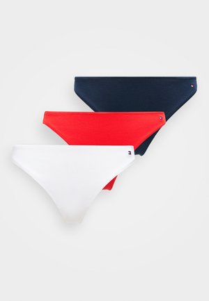 Giovane donna con capelli ricci che indossa un reggiseno bianco e slip bikini blu navy, in piedi vicino a una poltrona bianca testurizzata su uno sfondo semplice.