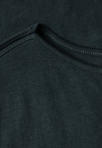 Tummanvihreä puuvillakangas, jossa on sileä pinta ja pyöreä pääntie. Näkyvä ommel kauluksen ja hartioiden ympärillä.