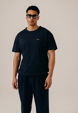 Ung mand iført sorte solbriller, en sort t-shirt med lille gult logo og sorte bukser, der står foran en ensfarvet beige baggrund.