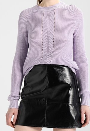 Femme portant un pull en maille violet clair et une mini-jupe en cuir noir brillant, debout avec les mains détendues le long du corps.