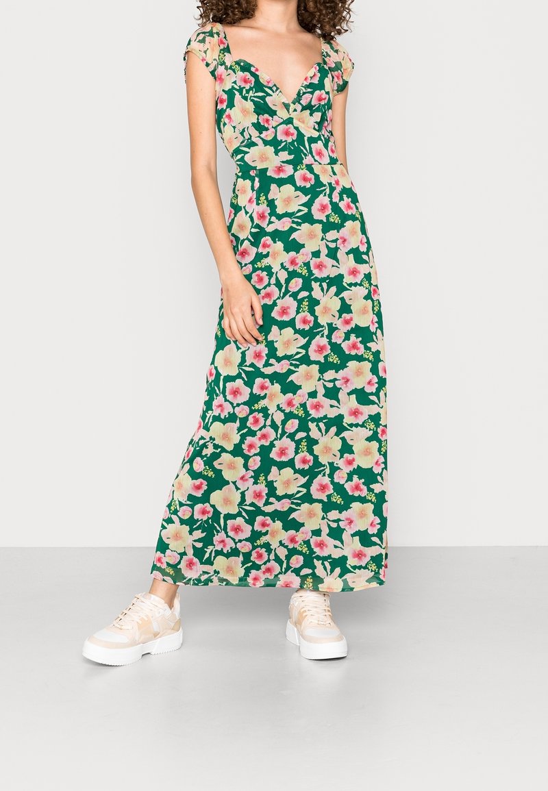 Blommig maxiklänning med djup V-ringning, korta ärmar i kappa, grön tyg med rosa och gula blommor, sammansatt med ljusa sneakers.