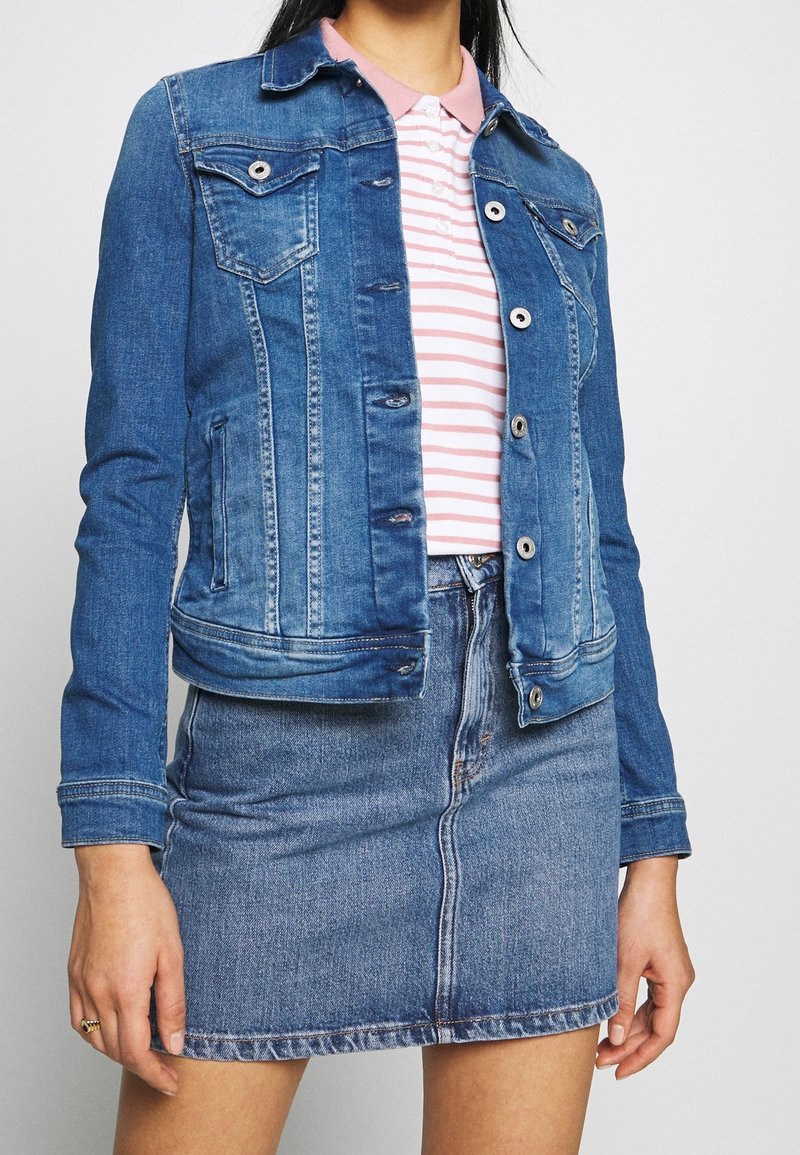 Chaqueta de denim con un lavado azul claro, dos bolsillos en el pecho y botones plateados, combinada con una polo a rayas rosa y blanca y una falda de denim.