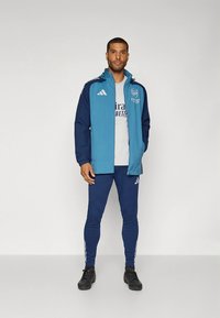 Urheilutakki teelinsinisessä ja navyssä, vetoketjulla ja logolla, yhdistetty navy-sävyisiin slim fit -housuihin. Mustat urheilukengät täydentävät asun.