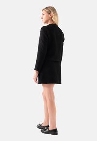 Zwarte textuurgeïnspireerde blazer met een slim fit ontwerp, gecombineerd met een bijpassende zwarte rok. Het model draagt glanzende zwarte schoenen met een strikaccent.