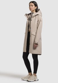 Manteau long beige imperméable avec une capuche, boutons noirs et deux poches avant. Assorti avec des leggings noirs et des baskets beiges avec des accents bleus.