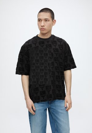 Νέος άντρας με κοντά μαλλιά που φοράει μαύρο μπλουζάκι με σχέδια και φαρδιά, ανοιχτόχρωμα τζιν, στέκεται μπροστά σε απλό λευκό φόντο.