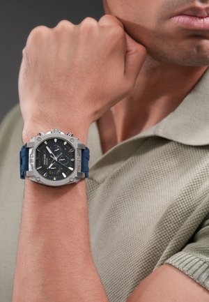 Man draagt een groen gebreid poloshirt en toont een polshorloge met een zilveren vierkante kast, een zwarte wijzerplaat en een blauwe band op een opgestoken vuist tegen een grijze achtergrond.