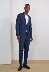 Traje azul marino de corte ajustado, con chaqueta de un solo botón y solapas en pico, combinado con una camiseta blanca y zapatos negros, de pie sobre un suelo de madera clara.
