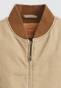 Beež jakk pruuni ribaga kraega, ees metallist tõmblukk ja Levi Strauss & Co silt krae sees.