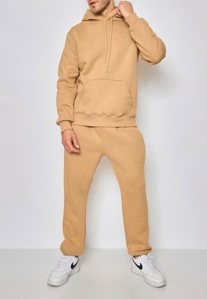 Homme portant un sweat à capuche beige et un pantalon de survêtement assorti avec des baskets blanches, debout devant un arrière-plan uni.