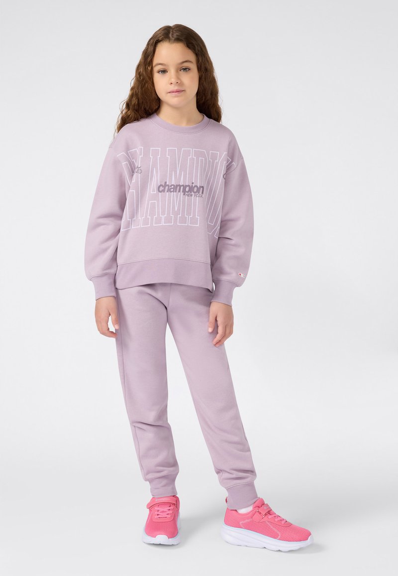 Sudadera lavanda con el logo de "Champion", combinada con joggers lavanda a juego y zapatillas rosas. Textura suave, diseño de ajuste casual.