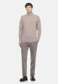 Uomo in piedi che indossa un maglione beige a collo alto, pantaloni grigi e scarpe eleganti nere su uno sfondo chiaro e semplice.