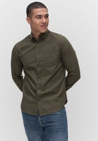 Olijfgroen hemd met lange mouwen, voorzien van een button-down kraag, een borstzak en bruine knopen, gecombineerd met blauwe denimjeans.