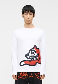 Νέος άνδρας που φοράει λευκό πουκάμισο με μακριά μανίκια και μαύρο παντελόνι, και τα δύο με μεγάλα κόκκινα και μαύρα ζωγραφισμένα ζωικά σχέδια.