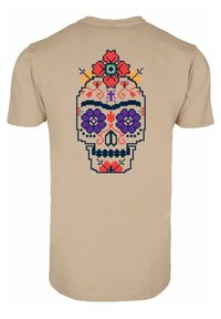 Beige t-skjorte med fargerik hodeskallegrafikk på baksiden, dekorert med blomsterdesign i lilla, rød og blå, med intrikate detaljer.