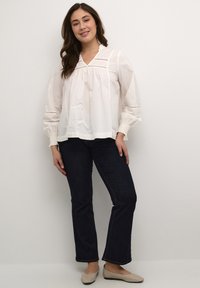Vit långärmad blus med hög halsringning, smockade manschetter och virkat detaljer. Matchad med mörka jeans och beige ballerinaskor.