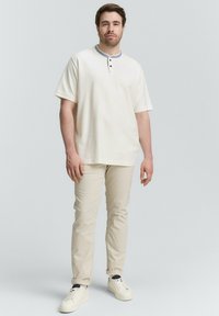 Mann steht in einem cremefarbenen Kurzarm-Henley-Shirt, beigen Hosen und weißen Sneakers vor einem schlichten hellen Hintergrund.