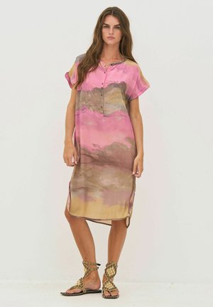 Nő áll, laza, rövid ujjú ruhát visel, amely rózsaszín, barna és sárga absztrakt mintákkal díszített, valamint bézs színű, szegecselt bokapántos szandált.