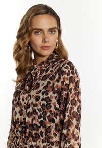 Blouse met luipaardprint in beige, bruin en zwart; heeft een rimpelkragen en lange mouwen. Glad materiaal met een vloeiende uitstraling.