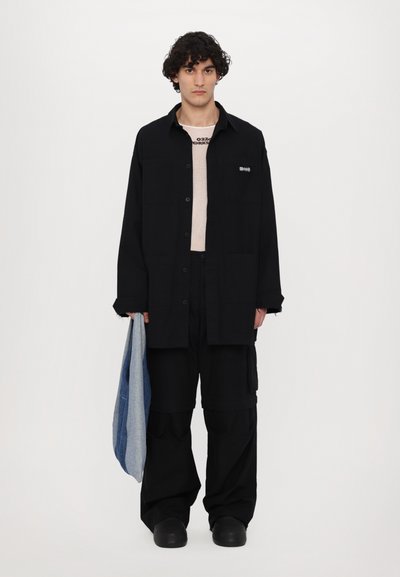Camisa negra de gran tamaño con cierre de botones, superpuesta sobre una camiseta beige clara. Pantalones cargo negros y sosteniendo una chaqueta de mezclilla doblada. Calzado negro.
