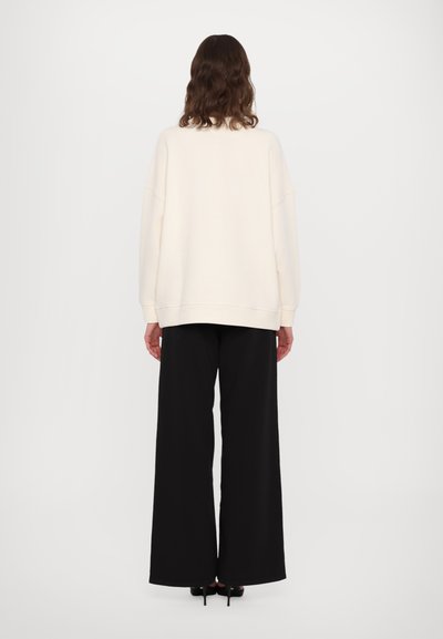Cremefarbenes oversized Sweatshirt mit gerippten Bündchen, kombiniert mit weit geschnittenen schwarzen Hosen, sichtbarem Bund und Schuhen mit Absätzen. Einfaches und unauffälliges Design.