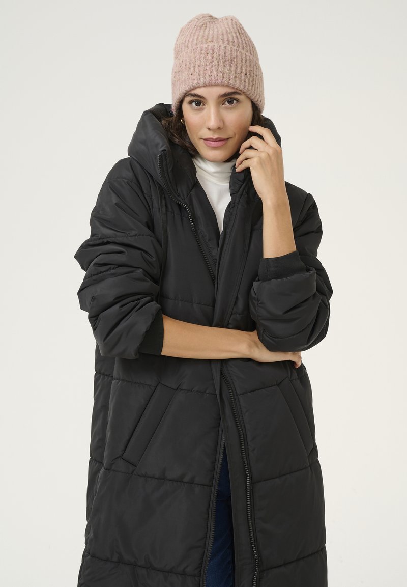 Black puffer coat with a zipper, showing quilted pattern, paired with a light pink ribbed knit beanie. Model is posing with arms crossed.