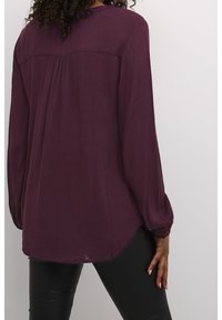 Langärmlige lila Bluse aus strukturiertem Stoff, mit yoke-Detail auf der Rückseite und abgerundetem Saum, getragen mit schwarzen, figurbetonten Hosen.