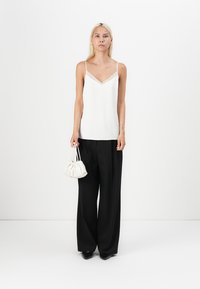 Weißes Camisole mit V-Ausschnitt und dünnen Trägern, kombiniert mit lockeren schwarzen Hosen und einer kleinen weißen Tasche mit Raffung, auf einem neutralen Hintergrund platziert.