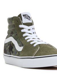 High-top tennarit, joissa on oliivinvihreää mokkaa ja camouflagemateriaalia, valkoiset nauhat, Vansin tunnuslogo ja valkoinen kumipohja.