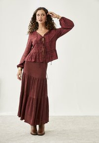 Blusa floral color burdeos con cordón, dobladillo peplum y mangas largas; falda maxi de capas a juego con botones. Botines marrones.