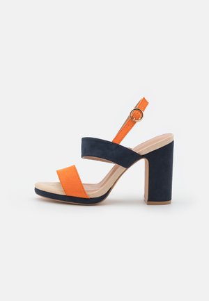 Sandal med høje hæle i navyblå med tyk hæl og stropper samt en kraftig orange frontstropp og ankelrem med spænde.