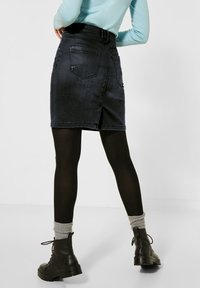 Falda de mezclilla negra con una cintura ajustada, bolsillos traseros y un dobladillo recto. Combinada con medias negras y botas gruesas de cordones con calcetas grises.