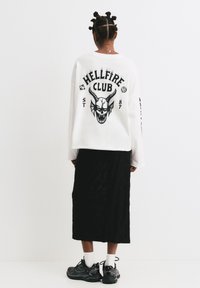Bílé oversize mikina s černým grafickým potiskem, na kterém je lebka a nápis „HELLFIRE CLUB“. V kombinaci s dlouhou černou texturovanou sukní a teniskami.