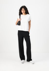 White short-sleeve t-shirt with printed branding, paired with black wide-leg trousers. Model holds a small black handbag and wears white sneakers.