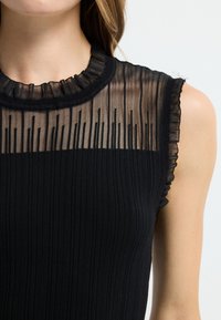 Czarny prążkowany top z przejrzystym, plisowanym dekoltem i subtelnymi falbankami. Materiał jest miękki, z dopasowanym kształtem i półprzezroczystą górną częścią.
