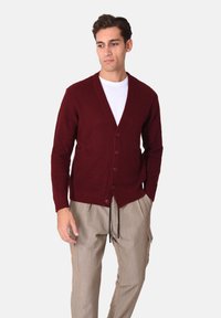 Cardigan bordeaux a coste con scollo a V, chiusura con bottoni e maniche lunghe, indossato sopra una camicia bianca, abbinato a pantaloni chiari.