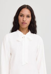 Chemise blanche à manches longues avec un col haut et une fermeture en nœud papillon. Tissu texturé avec un léger éclat et manches légèrement bouffantes.
