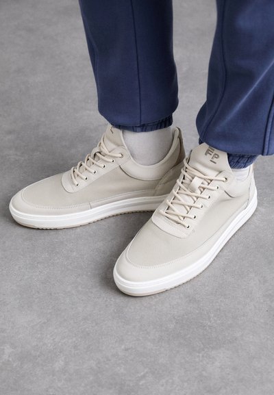Baskets beiges en matériau texturé, à bout rond, avec des lacets et un design minimaliste. Semelle blanche avec des détails beige clair. Porté avec un jogging bleu marine.