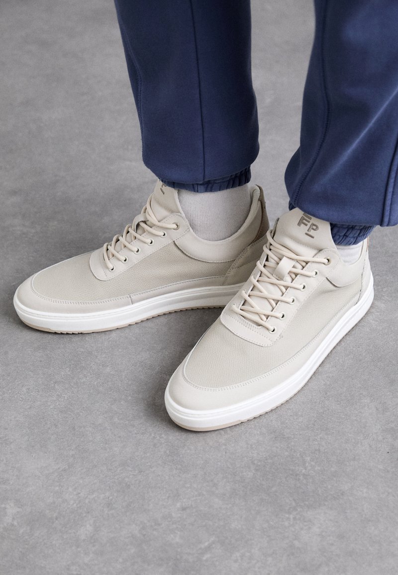 Beige sneaker van textielmateriaal, ronde neus, veters en een minimalistisch ontwerp. Witte zool met lichtbruine details. Draag met marineblauwe joggingbroeken.