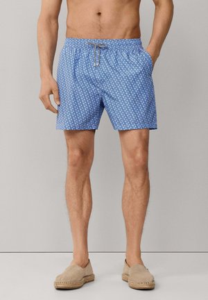 Homme portant un short de bain bleu à motifs et des espadrilles beige à enfiler, debout devant un fond gris clair uni.