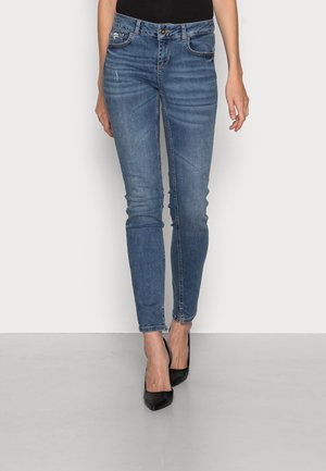 Slim fit blå jeans med let falmning og whiskering-detaljer, båret med sorte spidse hæle mod en ensfarvet baggrund.