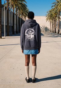 Personne portant un sweat à capuche foncé avec un motif floral et du texte, une jupe en jean, des chaussettes hautes jusqu'aux genoux et des chaussures noires, debout sur un chemin bordé de palmiers baigné de soleil.