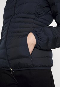 Laivastonsininen toppatakki, jossa on ommellut paneelit, vetoketjulliset taskut, joustavat hihansuut ja sileä kangaspinta. Etuvarmistuksessa näkyy vetoketju.