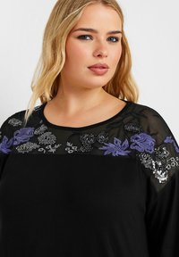 Schwarzes Oberteil mit floraler Stickerei in Lila und Silber auf einem transparenten Stoffyoke, mit rundem Halsausschnitt und lässigen Ärmeln.